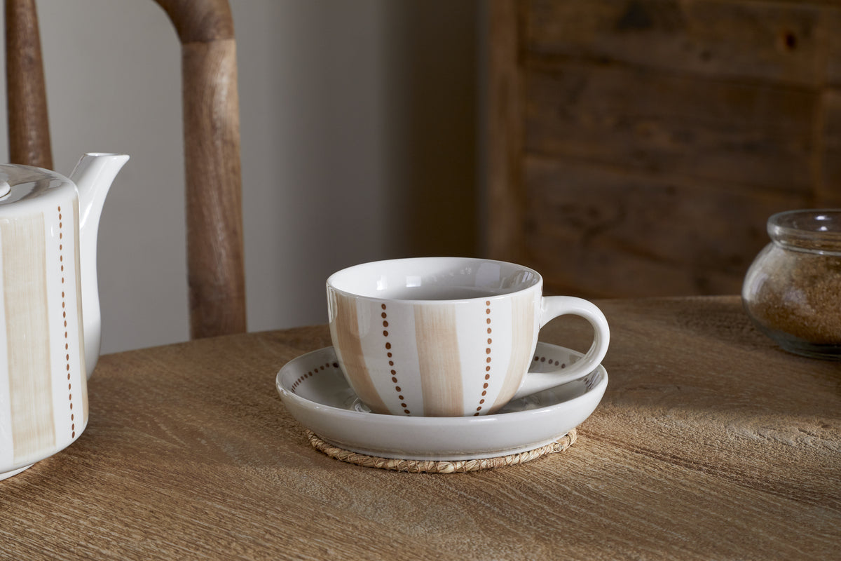 A nkuku Patthar Cup & Saucer - Cream & Clay with beige stripes rests on a wicker coaster, complemented by a matching teapot, glass jar of brown sugar, and a wooden chair and cabinet in the background.