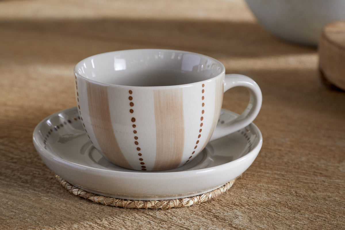The Patthar Cup & Saucer - Cream & Clay by nkuku features a handled cup on a matching saucer, both with beige stripes and dotted details, displayed on a woven coaster atop light wood, with soft neutral tones in the background.