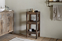 A rustic bathroom with wooden floors and light walls features the nkuku Akara Reclaimed Wood Storage Caddy - Natural, holding jars, a glass pitcher, and toothbrush cup near a towel rack with folded towels and a wooden vanity with sink and soap dispenser.