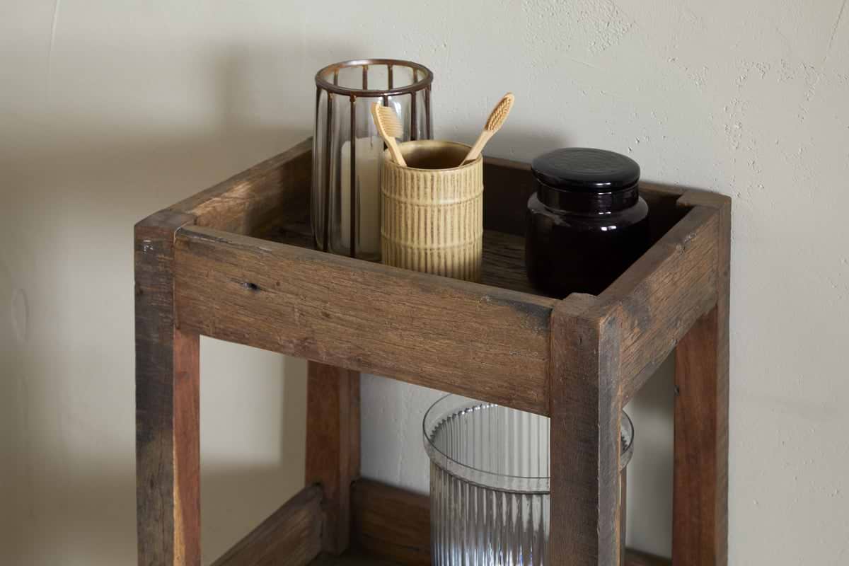 The Akara Reclaimed Wood Storage Caddy by nkuku holds a striped drinking glass with brown rim, a ribbed beige cup with bamboo toothbrushes, a dark glass jar, and a textured clear container on its shelves against a light backdrop.