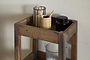 The Akara Reclaimed Wood Storage Caddy by nkuku holds a striped drinking glass with brown rim, a ribbed beige cup with bamboo toothbrushes, a dark glass jar, and a textured clear container on its shelves against a light backdrop.