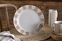 The nkuku Patthar Side Plate - Cream & Clay, with a wide rim and tan-cream geometric patterns, stands on a woven placemat beside matching tableware, a floral napkin, and rustic wooden table for a cozy touch.