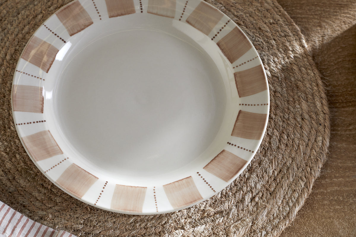 The nkuku Patthar Side Plate - Cream & Clay, featuring hand-painted beige and brown blocks with dotted rim details, rests on a woven placemat on a wooden table. A striped cloth appears in the bottom left corner.