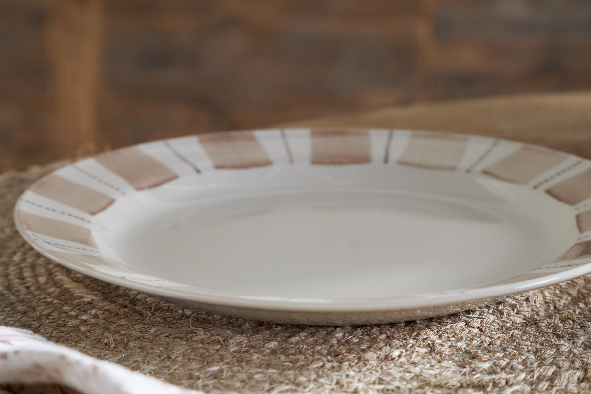 The Patthar Side Plate - Cream & Clay by nkuku, featuring a beige and white striped rim, rests on a woven placemat. Warm brown tones in the softly blurred background create a cozy vibe, while natural light highlights its elegant simplicity.