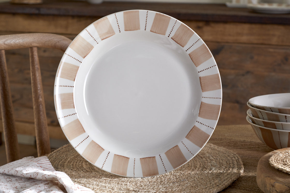 The nkuku Patthar Dinnerware Set (Cream & Clay, Set of 12) features round white ceramic plates with tan and beige borders, displayed on a woven placemat with matching hand-painted bowls stacked nearby. A wooden chair and rustic table complete the scene.
