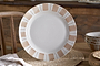 The Patthar Dinner Plate - Cream & Clay by nkuku, a hand-painted white ceramic with beige blocks and dotted brown lines on a wide rim, stands upright on a woven placemat, with matching bowls and rustic wooden furniture in the background.