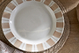 The Patthar Dinner Plate - Cream & Clay by nkuku, featuring a hand-painted rim of beige rectangles and dotted lines on white ceramic, sits empty on a woven placemat atop a wooden table beside striped cloth and bread.