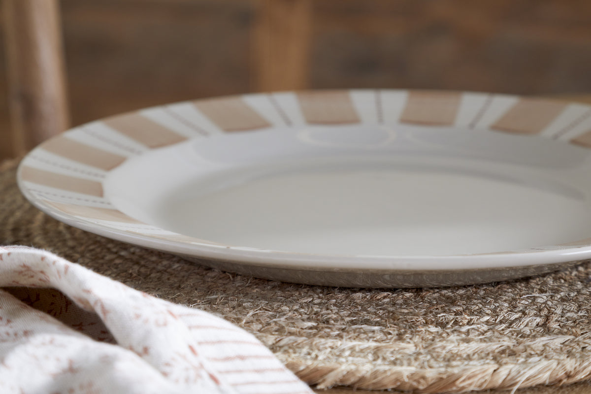 The nkuku Patthar Dinner Plate - Cream & Clay, featuring a shallow round design with a beige and white striped rim, rests on a woven placemat with a patterned cloth nearby and a blurred wooden background.