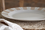 The nkuku Patthar Dinner Plate - Cream & Clay, featuring a shallow round design with a beige and white striped rim, rests on a woven placemat with a patterned cloth nearby and a blurred wooden background.