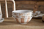 The Patthar Cereal Bowl - Cream & Clay by nkuku features hand-painted brown and beige striped and dotted designs. Stacked on a wooden table with rustic décor, these bowls add a cozy, homey touch to any dinnerware collection.