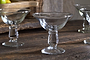The nkuku Sooraj Champagne Coupe set of 4 in clear glass features wide, shallow bowls and short stems. Displayed on a rustic wooden surface with fruit and a woven basket, these coupes exude timeless vintage charm.