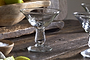 The Sooraj Champagne Coupe - Clear (Set of 4) by nkuku sits on a rustic wooden table with a bowl of green and yellow lemons, coarse salt, and a sliced lime, bringing vintage charm to your gathering.