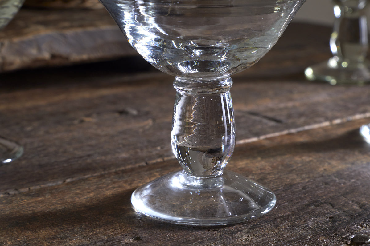 Close-up of the clear stem and base of an nkuku Sooraj Champagne Coupe (Set of 4), resting on a rustic wooden table with a softly blurred background that highlights the glass and wood textures.