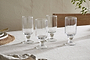 The Sooraj Large Glass - Clear (Set of 4) by nkuku, featuring short stems and elegant design, is displayed on a light textured tablecloth beside plates, cutlery, and a green plant stem.