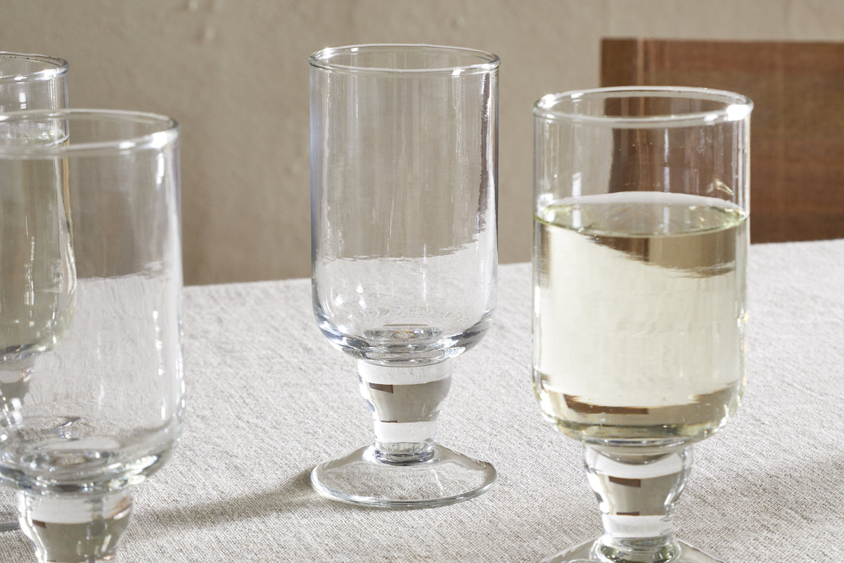 Four Sooraj Large Glass goblets by nkuku, from a set of 4, rest on a light tablecloth. Three are empty; the rightmost is half-filled with a pale summer drink. A simple, neutral background hints at a wooden chair.