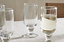 Four Sooraj Large Glass goblets by nkuku, from a set of 4, rest on a light tablecloth. Three are empty; the rightmost is half-filled with a pale summer drink. A simple, neutral background hints at a wooden chair.