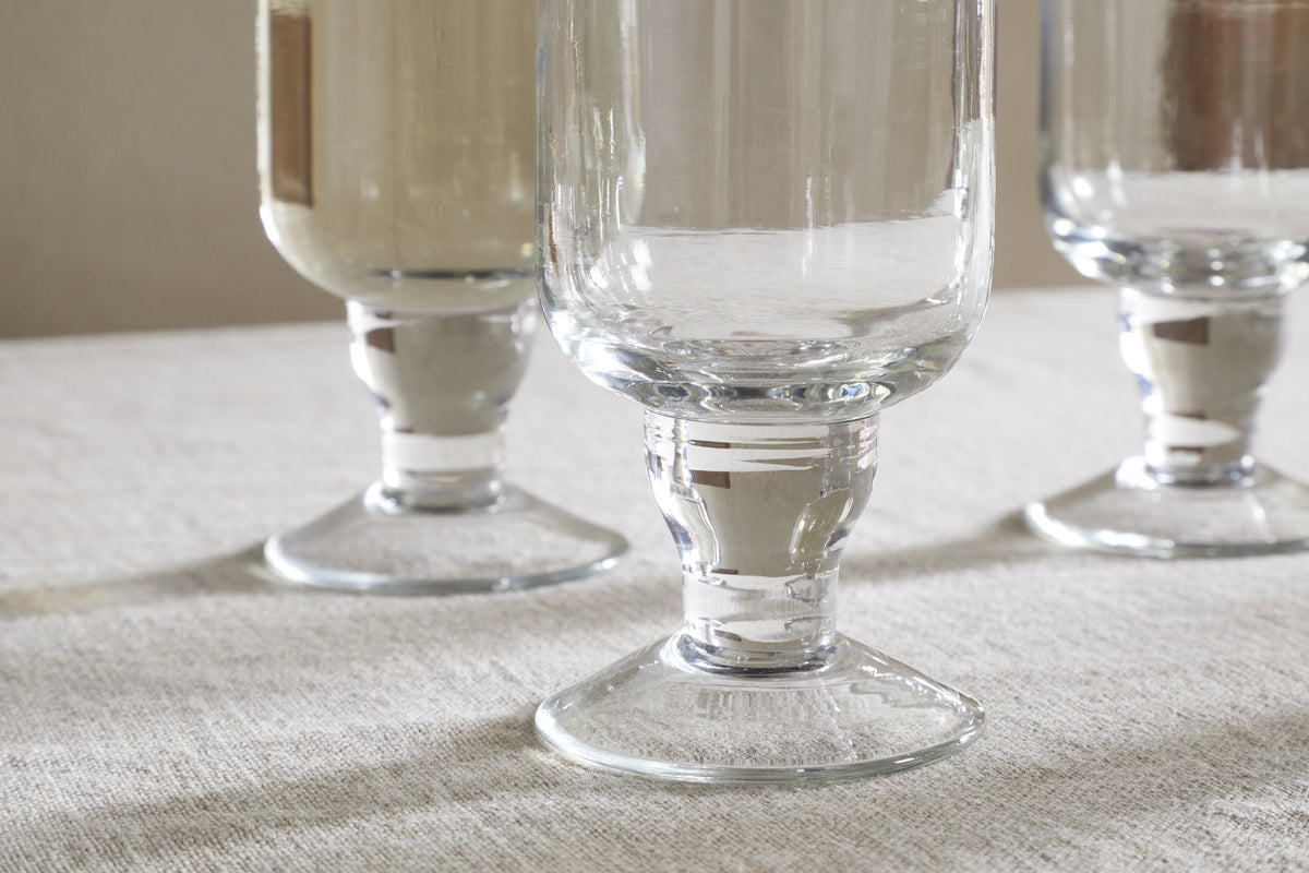 Three Sooraj Large Glasses by nkuku, from the clear glass set of 4, stand on a light-textured tablecloth. Each features a thick stem and wide base, showcasing stylish design and crystal-clear reflections.