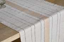A close-up of the nkuku Buna Table Runner - Clay, featuring natural stripes—thin black and one wide beige—draped over a wooden dining table, with a glass partially visible in the top left corner.