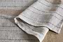 A close-up of the Buna Table Runner - Clay by nkuku, folded to reveal its textured chevron weave with natural stripes in thin navy blue and wider tan bands, draped softly on a rustic wooden surface.
