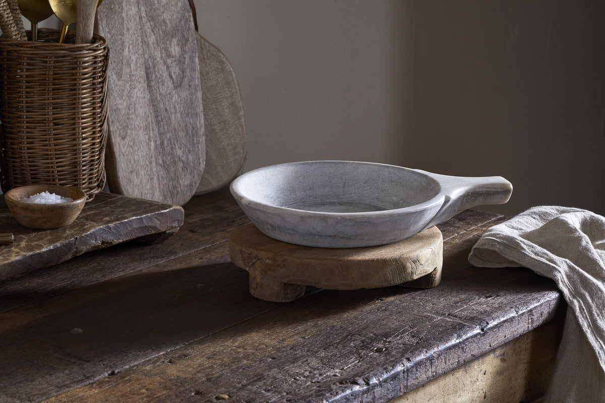 The nkuku Kaveeta Board - Natural sits on a rustic wooden trivet atop a weathered table, with its natural patina standing out. Nearby are a folded beige cloth, wicker utensil holder, cutting boards, and a bowl of coarse salt.