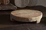 The Kaveeta Board - Natural by nkuku features a round, rustic wood design with visible grain and timeworn details, resting on a dark surface. Reclaimed boards and baskets in the background create an earthy, vintage feel.