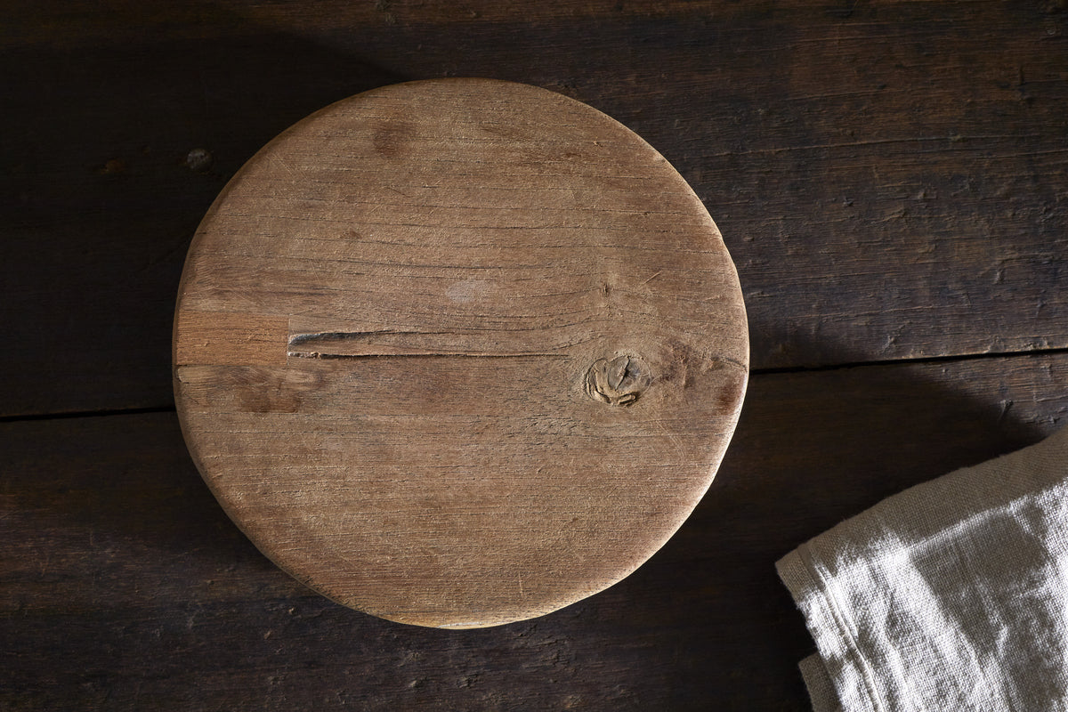 The Kaveeta Board - Natural by nkuku, a round reclaimed wood board with rustic grain, cracks, and knots, sits on a dark wood surface. A light beige textured cloth is visible in the image's bottom right corner.