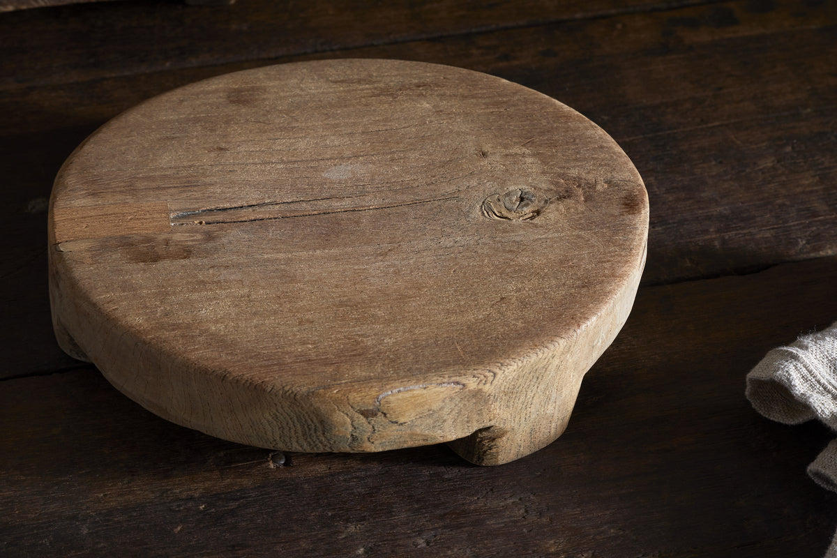 The nkuku Kaveeta Board - Natural, a round reclaimed wood board with rustic patina and timeworn charm, rests on a dark surface beside a folded light cloth, creating a warm, vintage kitchen vibe.