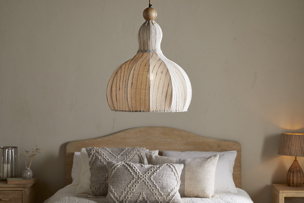 The nkuku Kela Fabric Pendant in Off White hangs above a wooden bed with a curved headboard, neatly dressed in neutral pillows and a white blanket, creating a calming ambience against the plain beige wall.