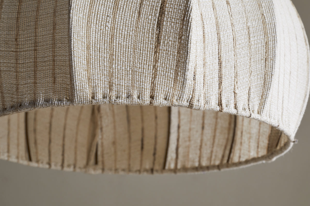 The nkuku Kela Fabric Pendant in off white features a woven linen shade with slim vertical brown lines. Its gentle curve and visible stitching showcase artisanal craftsmanship, bringing a calming ambience to your space.