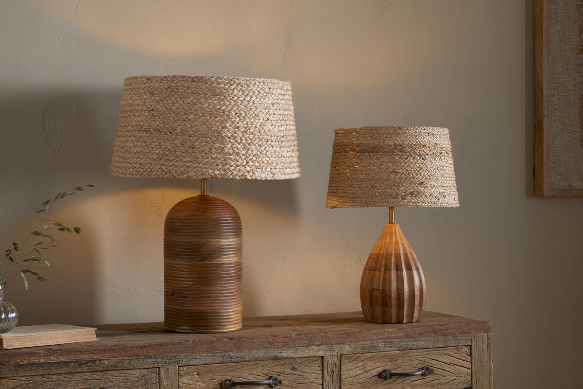 Two nkuku table lamps with Pisang Natural Lampshades sit on a rustic dresser, adding organic texture and warm light. The taller has a rounded dark brown base, while the shorter features a lighter, teardrop-shaped wooden base.