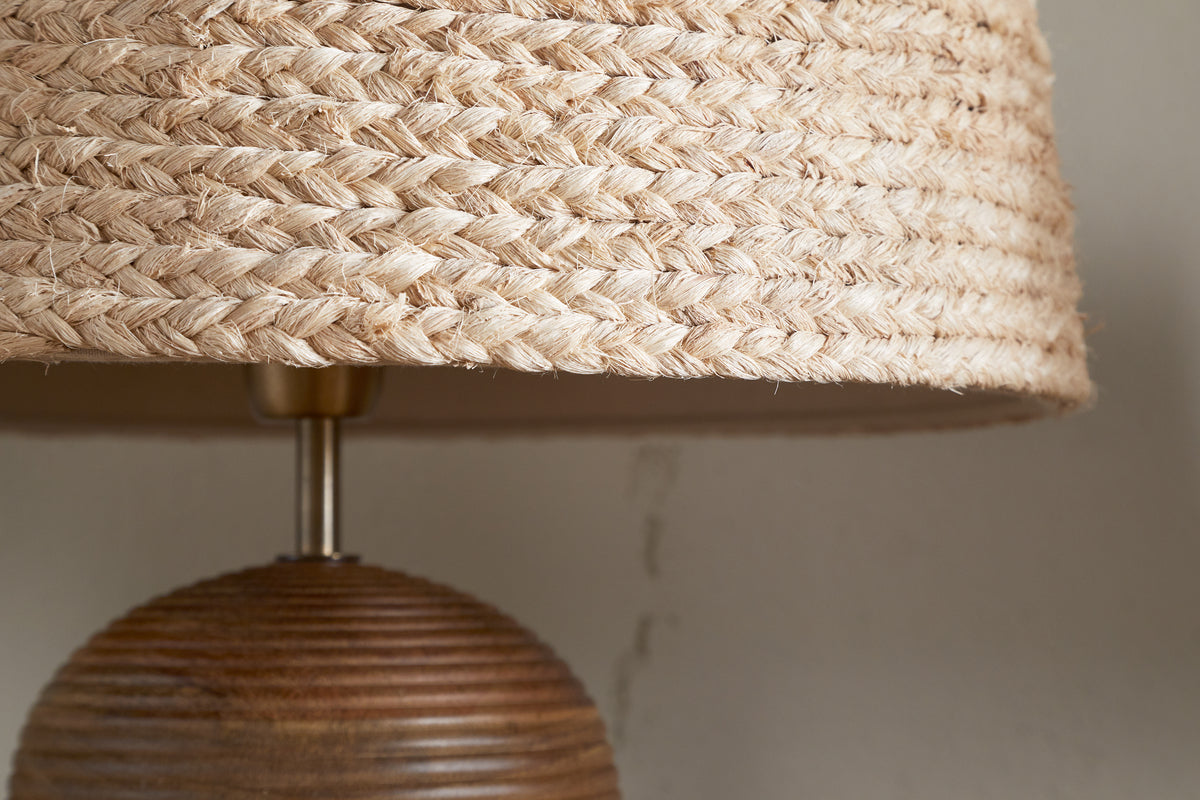 Close-up of the nkuku Pisang Natural Lampshade - Natural, featuring a woven jute shade and ribbed wooden base, set against a light-colored wall with a visible crack.