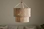 The Thimba Woven Jute Chandelier by nkuku, featuring a double-layered natural jute design, hangs above a neutral couch with pillows, casting a soft glow and rustic texture from matching woven cords.