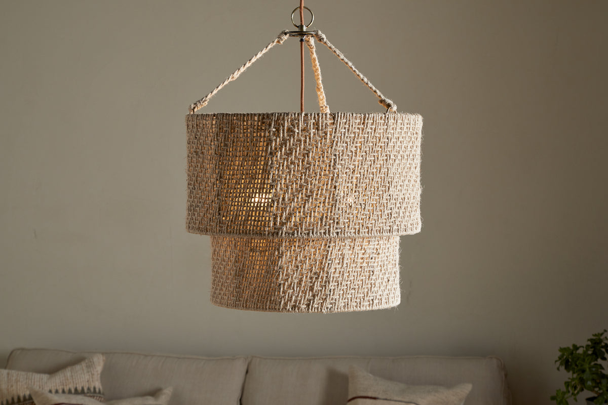 The Thimba Woven Jute Chandelier by nkuku, featuring a double-layered natural jute design, hangs above a neutral couch with pillows, casting a soft glow and rustic texture from matching woven cords.