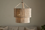 The Thimba Woven Jute Chandelier by nkuku, featuring a double-layered natural jute design, hangs above a neutral couch with pillows, casting a soft glow and rustic texture from matching woven cords.