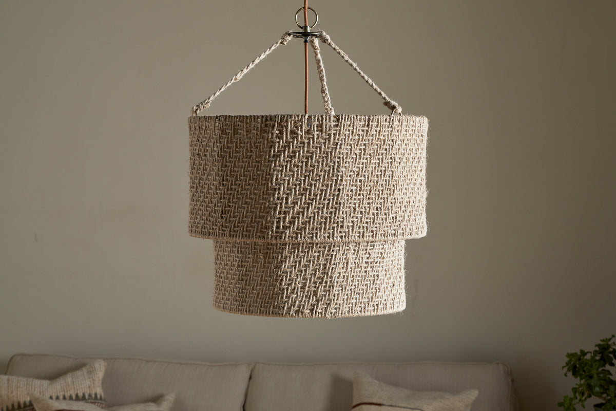 The Thimba Woven Jute Chandelier by nkuku hangs from the ceiling against a plain, light wall. Below, a neutral sofa with textured pillows adds rustic coziness and minimalist charm to the room.