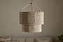 The Thimba Woven Jute Chandelier by nkuku hangs from the ceiling against a plain, light wall. Below, a neutral sofa with textured pillows adds rustic coziness and minimalist charm to the room.