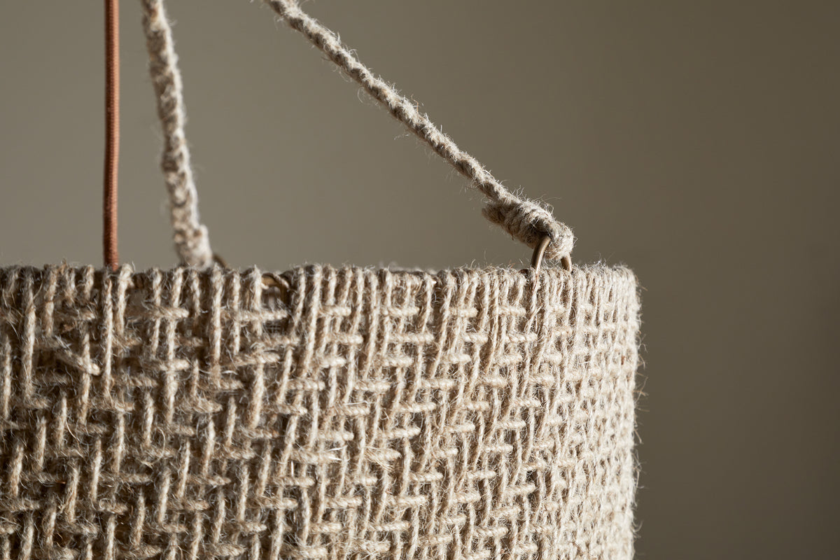Close-up of the Thimba Woven Jute Chandelier by nkuku, showcasing its rustic texture, zig-zag pattern, and thick rope handle threaded through a metal ring. Set against a neutral beige background with gentle natural light.
