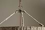 Close-up of the Thimba Woven Jute Chandelier - Natural by nkuku, featuring a rustic natural fiber shade and three braided ropes connecting to a metal ring, suspended from a cord against a plain beige background.