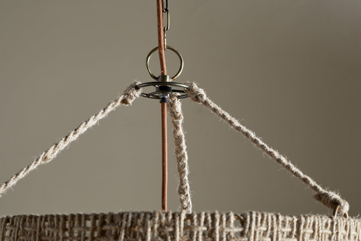 Close-up of the Thimba Woven Jute Chandelier - Natural by nkuku, featuring a rustic natural fiber shade and three braided ropes connecting to a metal ring, suspended from a cord against a plain beige background.