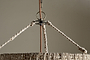 Close-up of the Thimba Woven Jute Chandelier - Natural by nkuku, featuring a rustic natural fiber shade and three braided ropes connecting to a metal ring, suspended from a cord against a plain beige background.