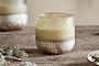 The nkuku Oakmoss & Rose 1 Wick Scented Candle - Yellow features creamy yellow wax in a hand-blown glass holder with earthy brown and off-white tones, adding a natural, cozy touch to any rustic wooden surface.