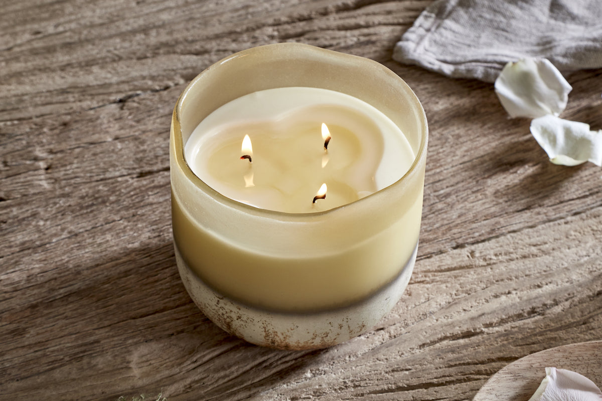 The nkuku Oakmoss & Rose 3 Wick Scented Candle - Yellow glows in a rustic, round two-toned holder on textured wood, with silk petals and a light gray cloth nearby for a cozy, serene decor touch.
