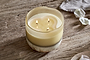 The nkuku Oakmoss & Rose 3 Wick Scented Candle - Yellow glows in a rustic, round two-toned holder on textured wood, with silk petals and a light gray cloth nearby for a cozy, serene decor touch.