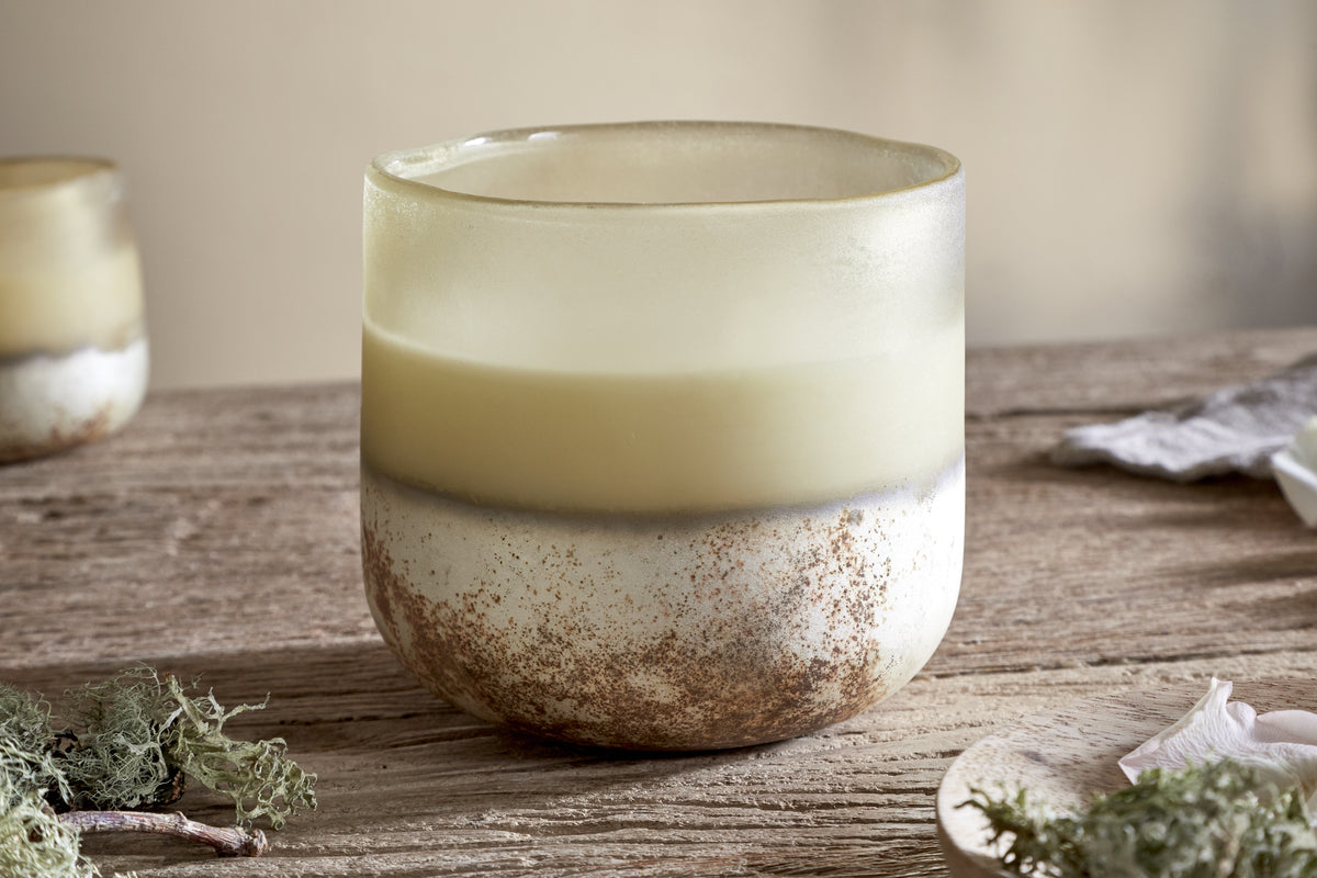 The nkuku Oakmoss & Rose 3 Wick Scented Candle in yellow sits on a rustic wood surface, accented by green moss and wood shavings. Its hand-blown glass vessel and earthy tones give it natural charm against a softly blurred background.