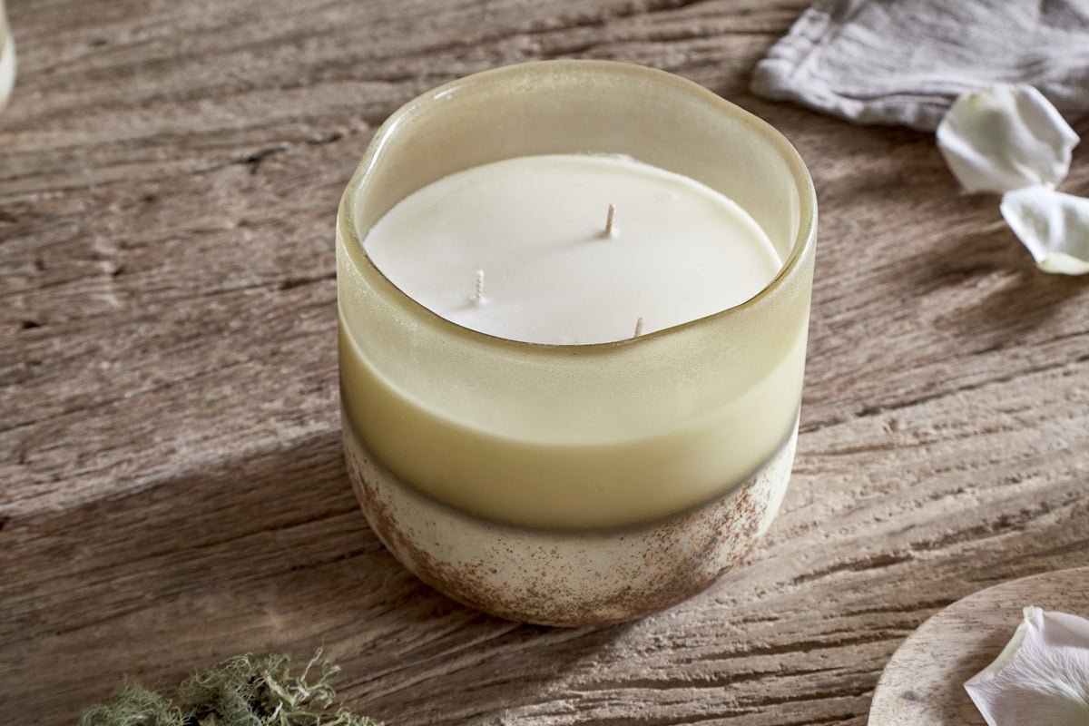The nkuku Oakmoss & Rose 3 Wick Scented Candle in yellow sits in a frosted, hand-blown glass vessel with beige and earthy tones on a rustic wooden surface, beside a cloth and delicate white flower petals.