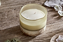 The nkuku Oakmoss & Rose 3 Wick Scented Candle in yellow sits in a frosted, hand-blown glass vessel with beige and earthy tones on a rustic wooden surface, beside a cloth and delicate white flower petals.