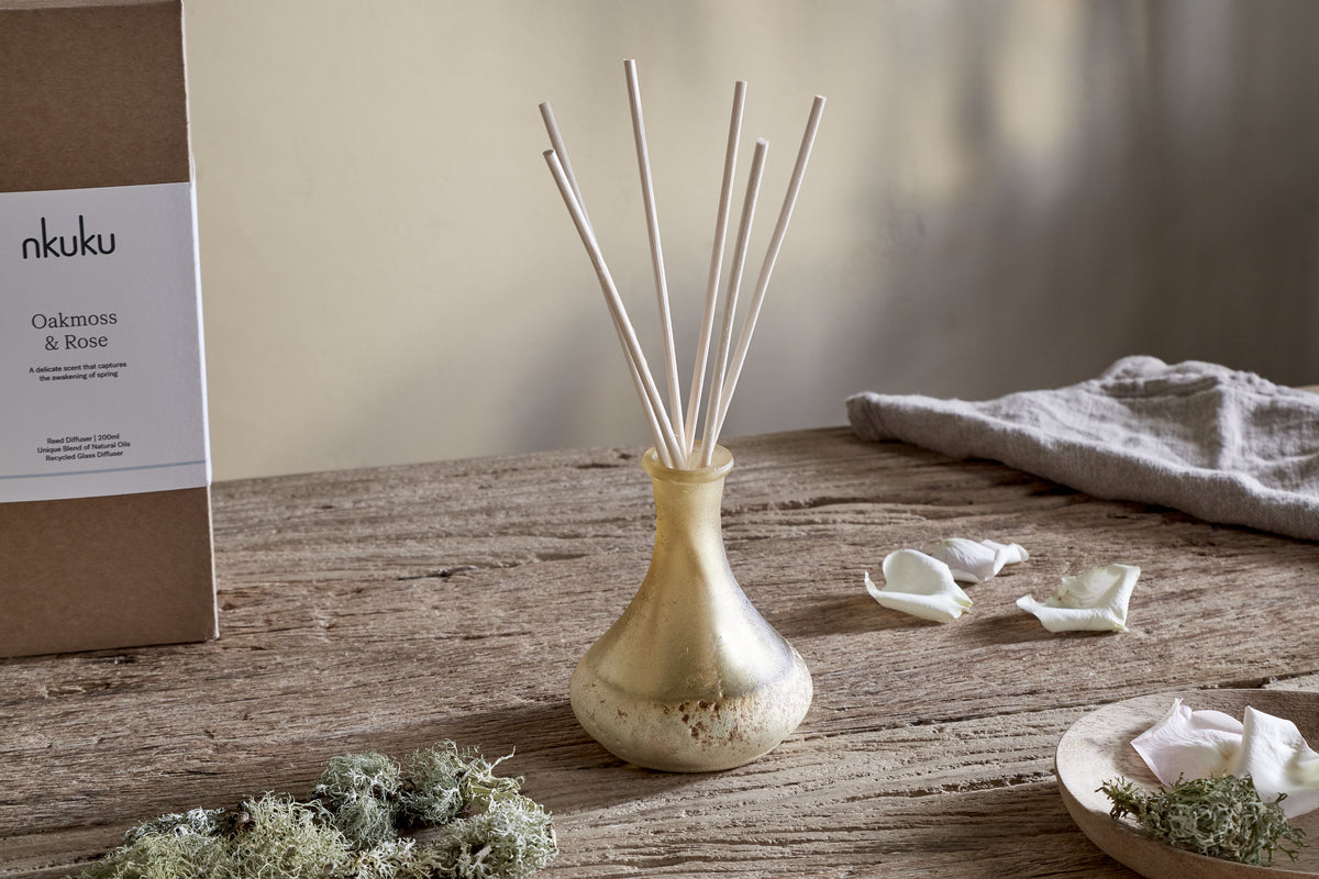 A small yellow nkuku Oakmoss & Rose Scented Reed Diffuser with white reeds sits on a rustic wooden table, surrounded by green moss, white rose petals, and its brown box, creating an inviting floral woody atmosphere.