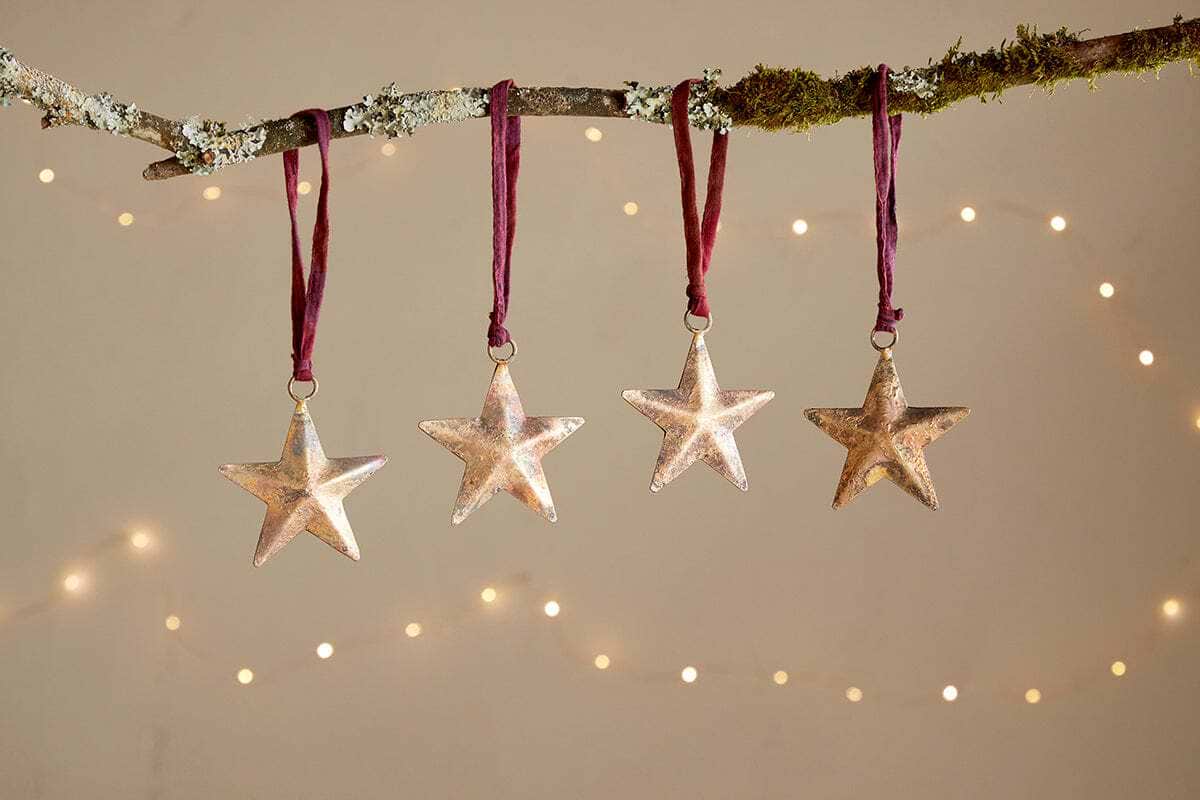 Four Roshani Star Baubles by nkuku, finished in antique brass, hang from burgundy ribbons on a mossy branch. Soft fairy lights blur behind, adding festive elegance and minimalist charm. Set of 4.