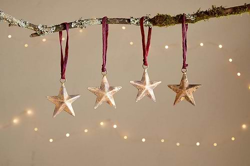 Four Roshani Star Baubles by nkuku, finished in antique brass, hang from burgundy ribbons on a mossy branch. Soft fairy lights blur behind, adding festive elegance and minimalist charm. Set of 4.