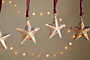 The nkuku Roshani Star Baubles - Antique Brass (Set of 4) hang from red strings against a beige backdrop with soft string lights. Only parts of the two outer star silhouettes are visible at the edges, creating a warm, festive feel.